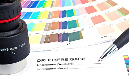 Nahaufnahme von Farbmusterkarten, Lupe und Kugelschreiber auf einem Druckfreigabe-Formular.